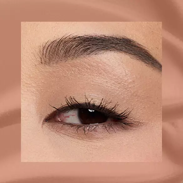 Close-up of an eye area with NYX Professional Makeup Lid Lingerie Cream Eye Pigment in Beige Appeal, shown on a tan matte textured background.