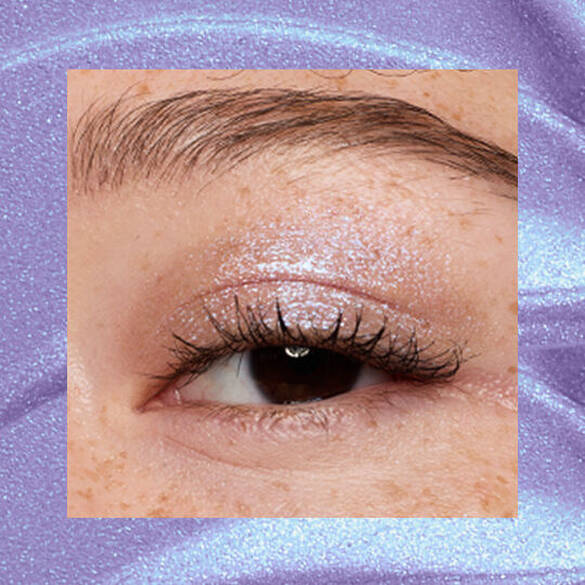 Close-up of an eye area with NYX Professional Makeup Lid Lingerie Cream Eye Pigment in Lacy Lilac, shown on a duochrome blue textured background.