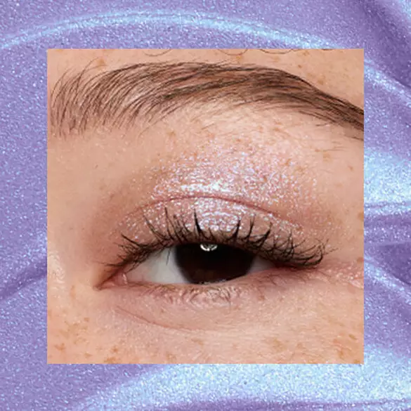 Close-up of an eye area with NYX Professional Makeup Lid Lingerie Cream Eye Pigment in Lacy Lilac, shown on a duochrome blue textured background.