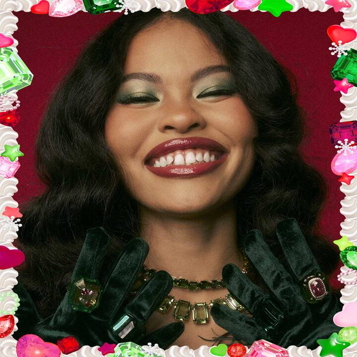 Visuel de campagne Halloween pour NYX Professional Makeup montrant un mannequin portant un maquillage felin rose et blanc, des gants en satin rose et tenant un tube de Butter Gloss rose metallise avec des paillettes. Il y a un fond de feuillage tropical vert.