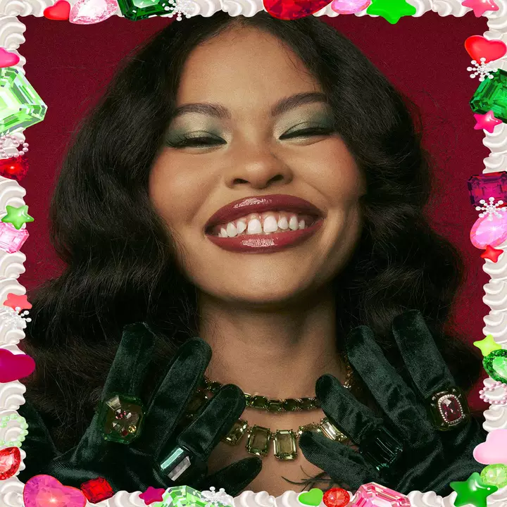 Visuel de campagne Halloween pour NYX Professional Makeup montrant un mannequin portant un maquillage felin rose et blanc, des gants en satin rose et tenant un tube de Butter Gloss rose metallise avec des paillettes. Il y a un fond de feuillage tropical vert.