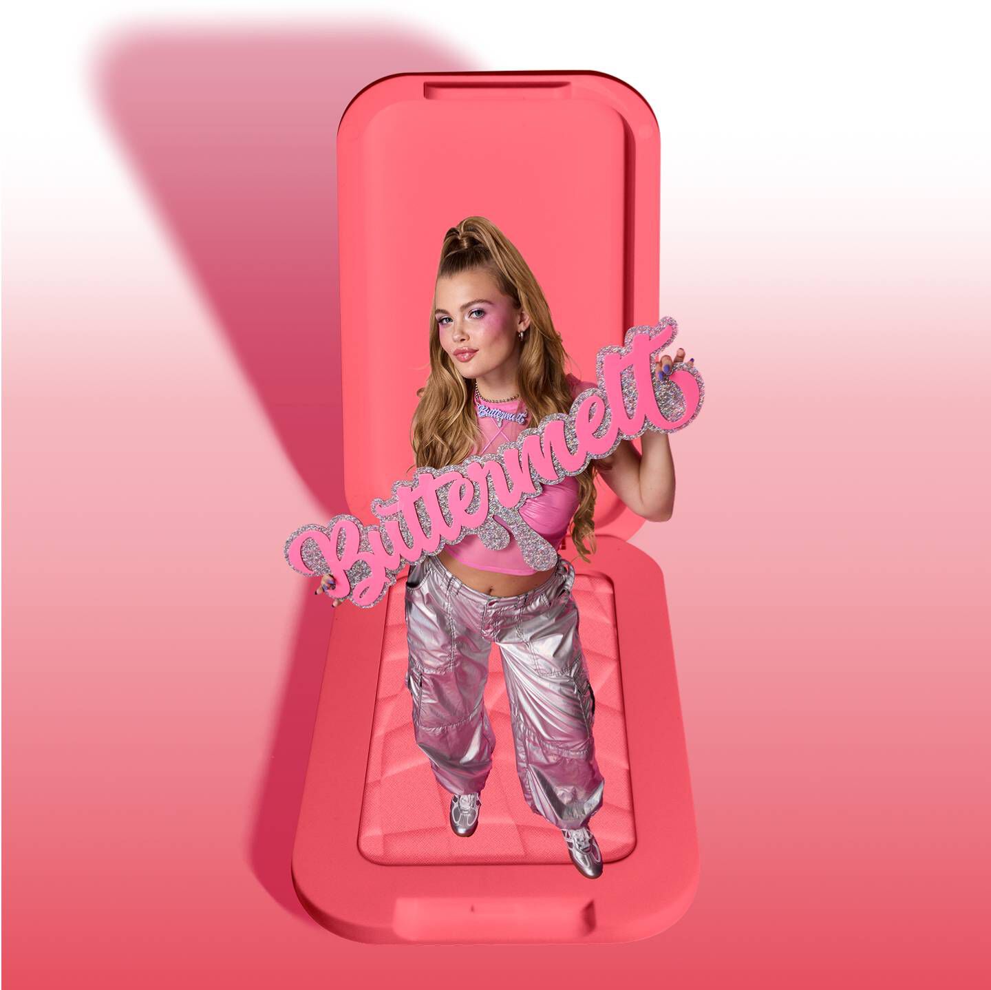 Photo d'une jeune femme aux longs cheveux blonds, vetue d'un haut rose et d'un pantalon cargo argente, tenant une decoupe Buttermelt rose pailletee. Elle se tient debout sur un grand poudrier de blush rose ouvert. L'arriere-plan presente un degrade de rose. Plan a hauteur des yeux, plan moyen, eclairage artificiel vif.