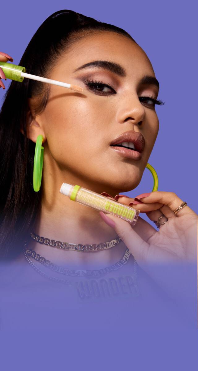 Sur un fond violet, une mannequin tient un produit dans une main et un pinceau dans l'autre au niveau de sa joue. On peut lire "WONDER SNATCH" en vert sur le produit. Sur le cote des produits sont regroupes au milieu a droite de l'image, il y a un autre anticernes WONDER SNATCH, des poudres fixatrices roses ouvertes, des fonds de teint MakeEm Wonder.