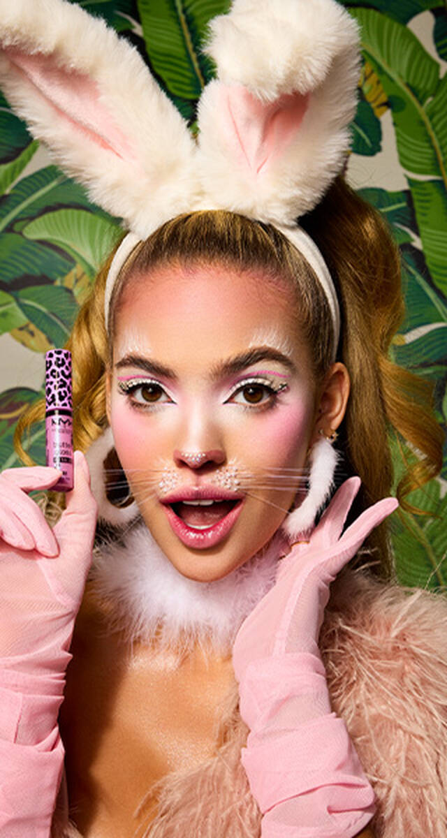 "Visuel de campagne Halloween pour NYX Professional Makeup montrant un mannequin portant un maquillage felin rose et blanc, des gants en satin rose et tenant un tube de Butter Gloss rose metallise avec des paillettes. Il y a un fond de feuillage tropical vert.